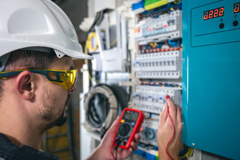 An electrical technician in safety gear works on a switchboard with fuses, inspecting or repairing electrical components.