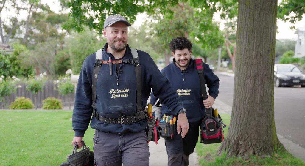 Two Statewide Sparkies electricians in branded uniforms and tool belts smiling while walking along a suburban Melbourne street