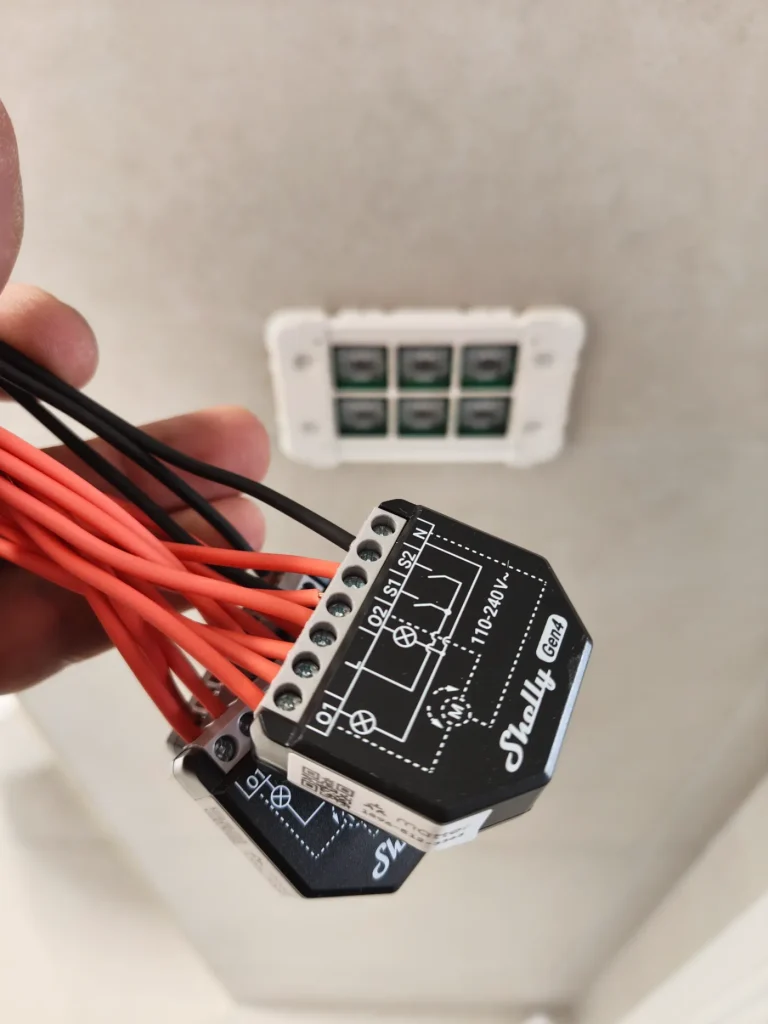 Electrician holding Shelly smart relay modules with wiring during a home automation installation, with a smart wall switch in the background